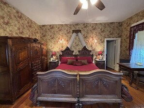 A bedroom with a large bed, wooden wardrobe, two bedside tables with lamps, floral wallpaper, and a ceiling fan.