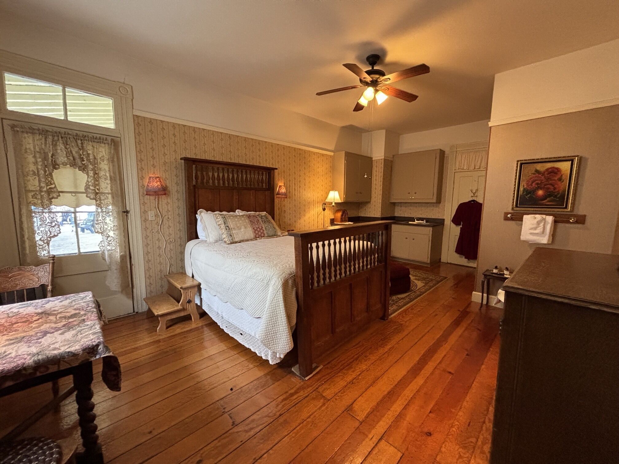 A bedroom with a wooden bed, a ceiling fan, a nightstand, a dresser, a window with curtains, and a painting on the wall.