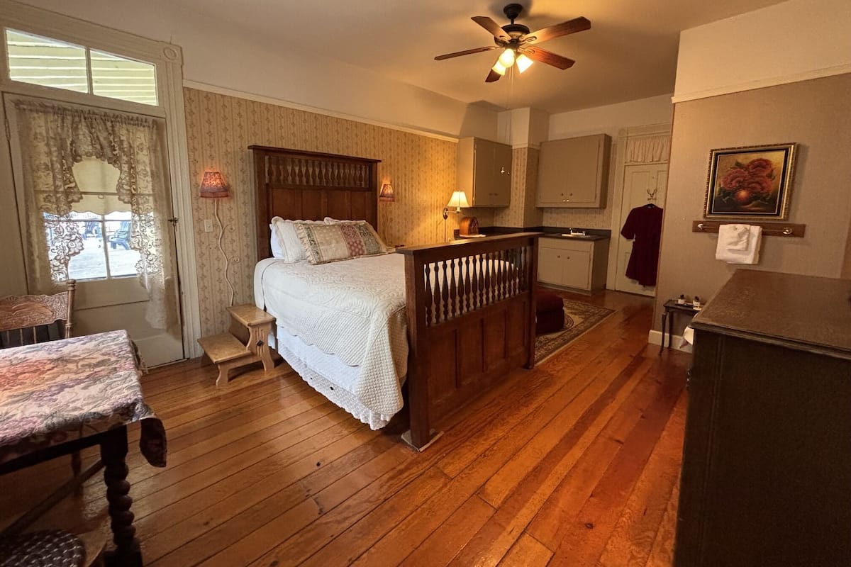 Un dormitorio con cama de madera, ventilador de techo, mesita de noche, cómoda, ventana con cortinas y un cuadro en la pared.