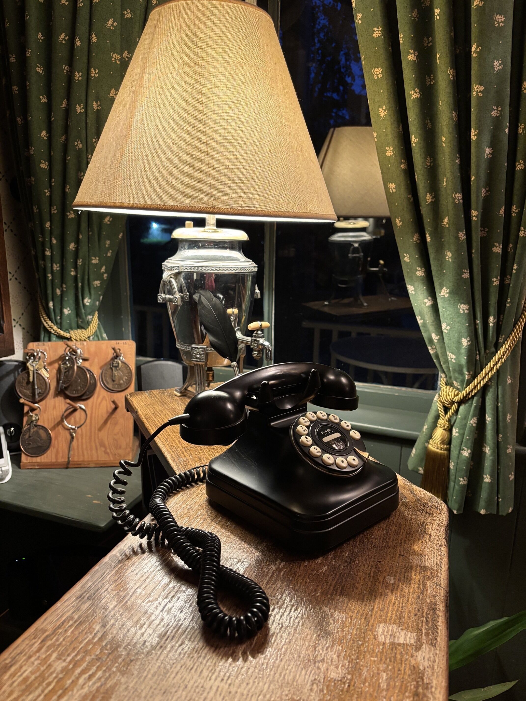 Un vieux téléphone noir à cadran sur un bureau en bois, une lampe avec un abat-jour beige et une fenêtre avec des rideaux verts.