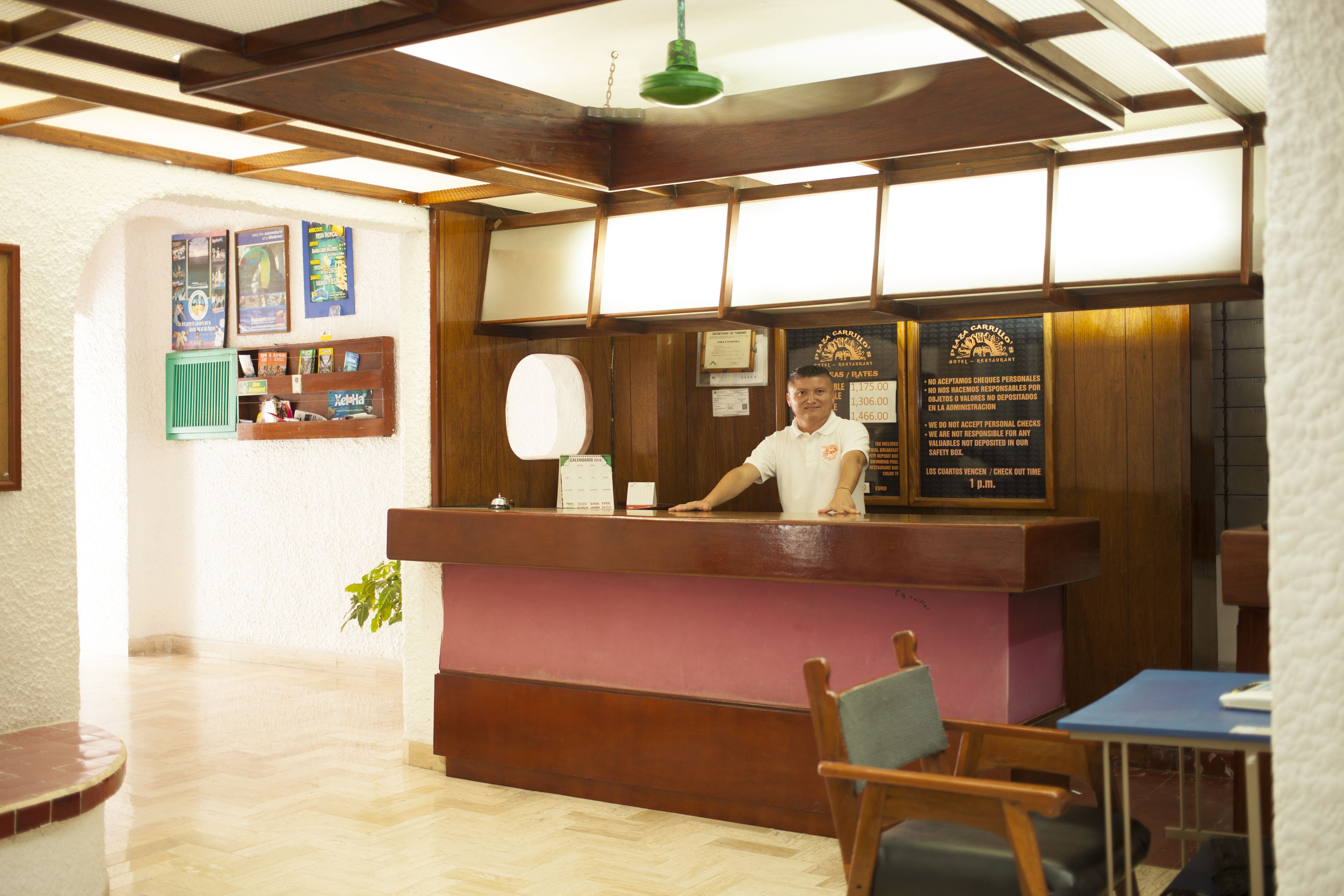 A reception desk with a man standing behind it, a wooden counter, a green plant, and a sign with text.