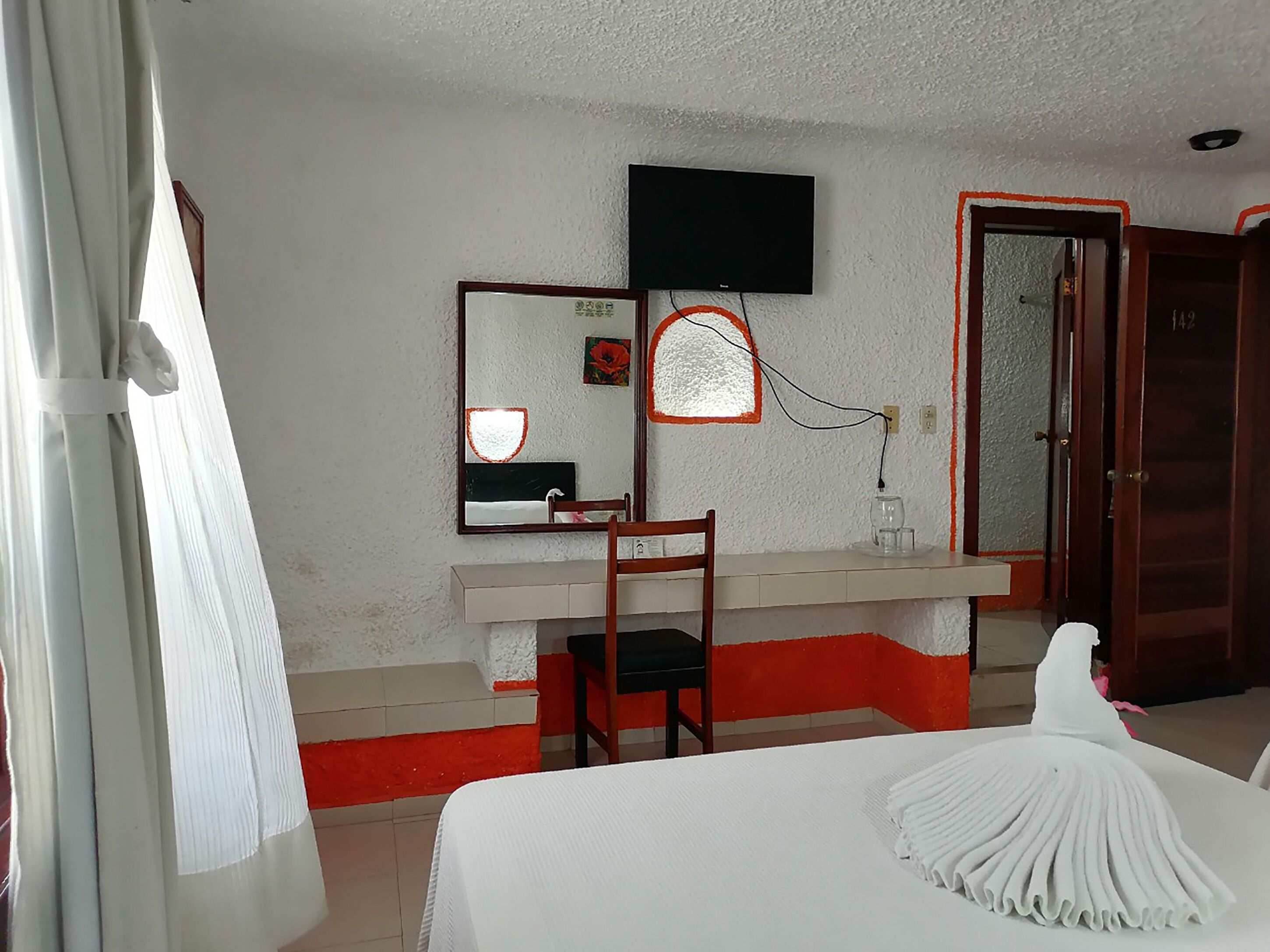 Una habitación de hotel con cama, escritorio, silla, televisor montado en la pared y una puerta.