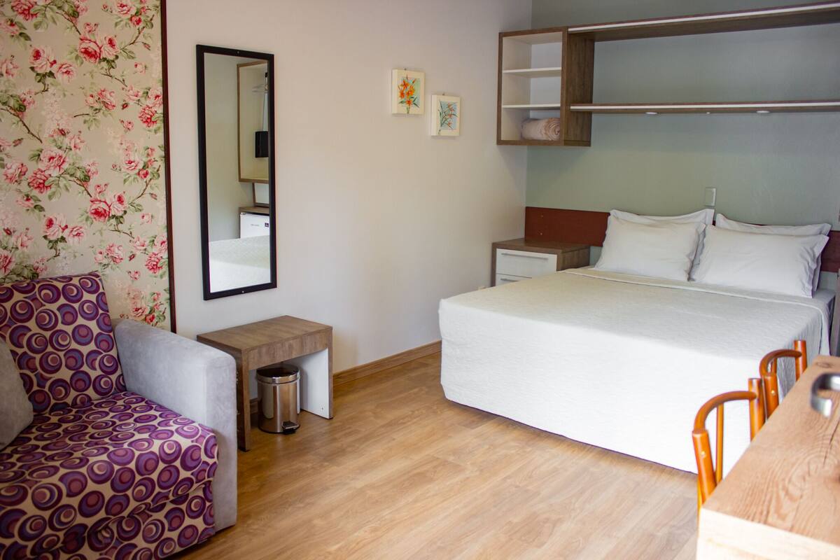 A hotel room with a bed, a wooden nightstand, a small table, a trash bin, a chair, and a sofa.