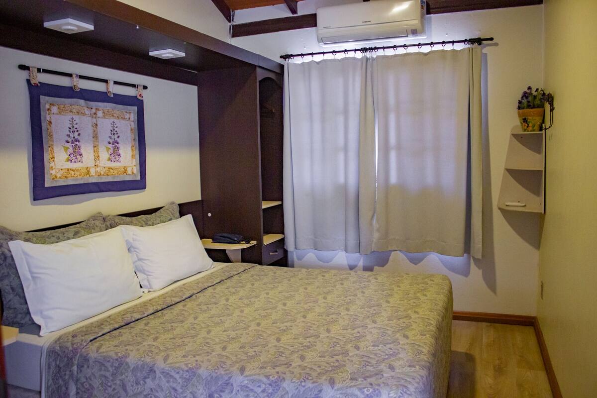 A bedroom with a bed, a nightstand, a wall-mounted air conditioner, a curtain, and a shelf with a vase.