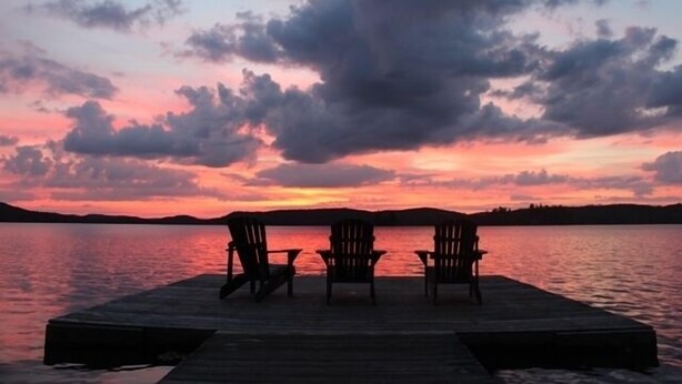 Un muelle de madera con tres sillas Adirondack mirando una puesta de sol sobre un lago tranquilo.