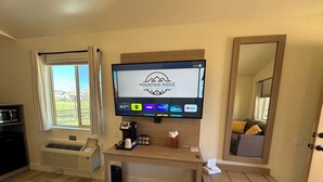 A hotel room with a large flat-screen TV displaying the Mountain Ridge logo, a wooden desk, a chair, a bed, a window with curtains, a microwave, a coffee maker, and a thermostat. - Mountain Ridge Cabins and Lodging (Hatch)