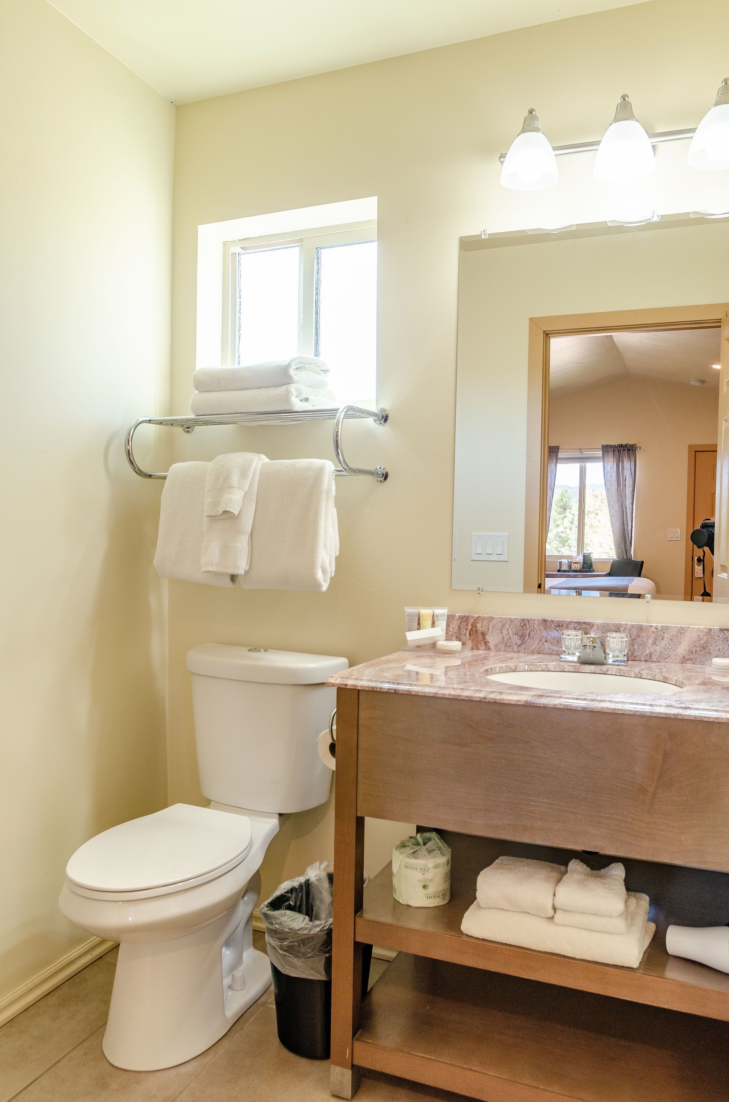 A bathroom with a toilet, sink, and mirror. There are towels on a rack and a window above the toilet.