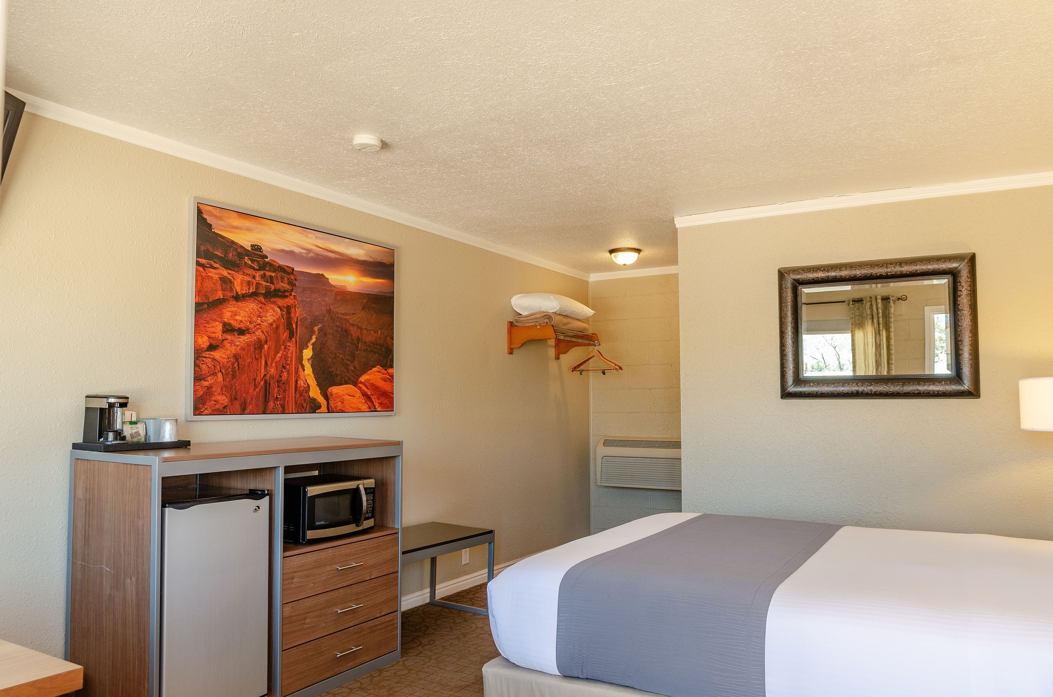 Ein Hotelzimmer mit einem Bett, einem Schreibtisch mit Mikrowelle, einem kleinen Kühlschrank, einem Canyonbild, einem hängenden Handtuchhalter und einem Spiegel an der Wand.