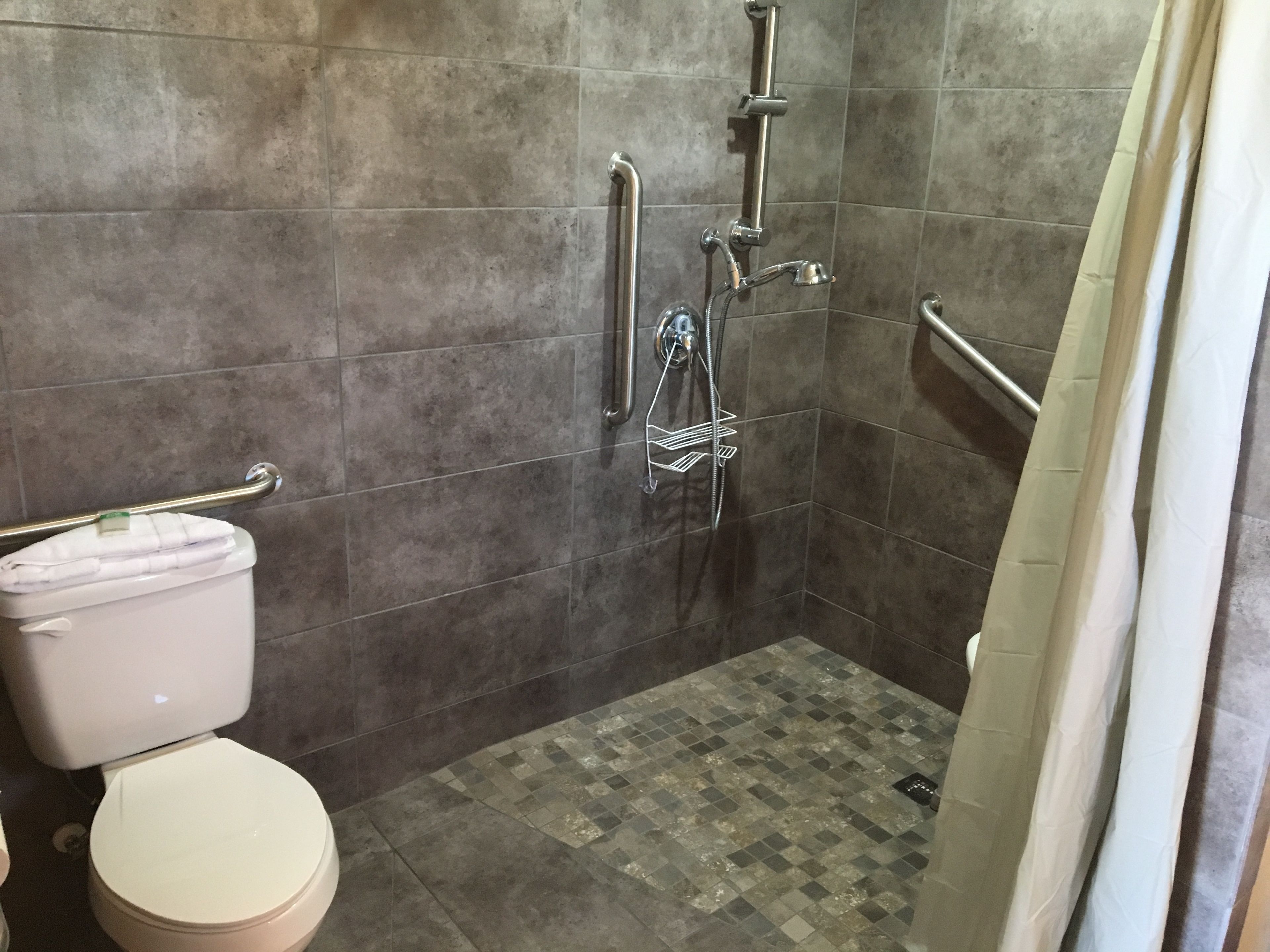 Un baño con inodoro, ducha con barra de apoyo y suelo de baldosas.