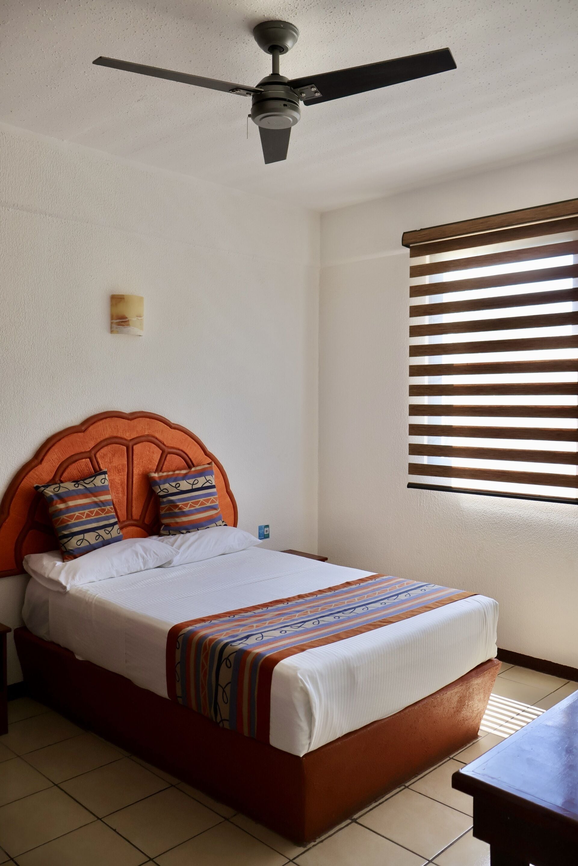 A neatly made bed with a wooden headboard, a ceiling fan, and a window with blinds.