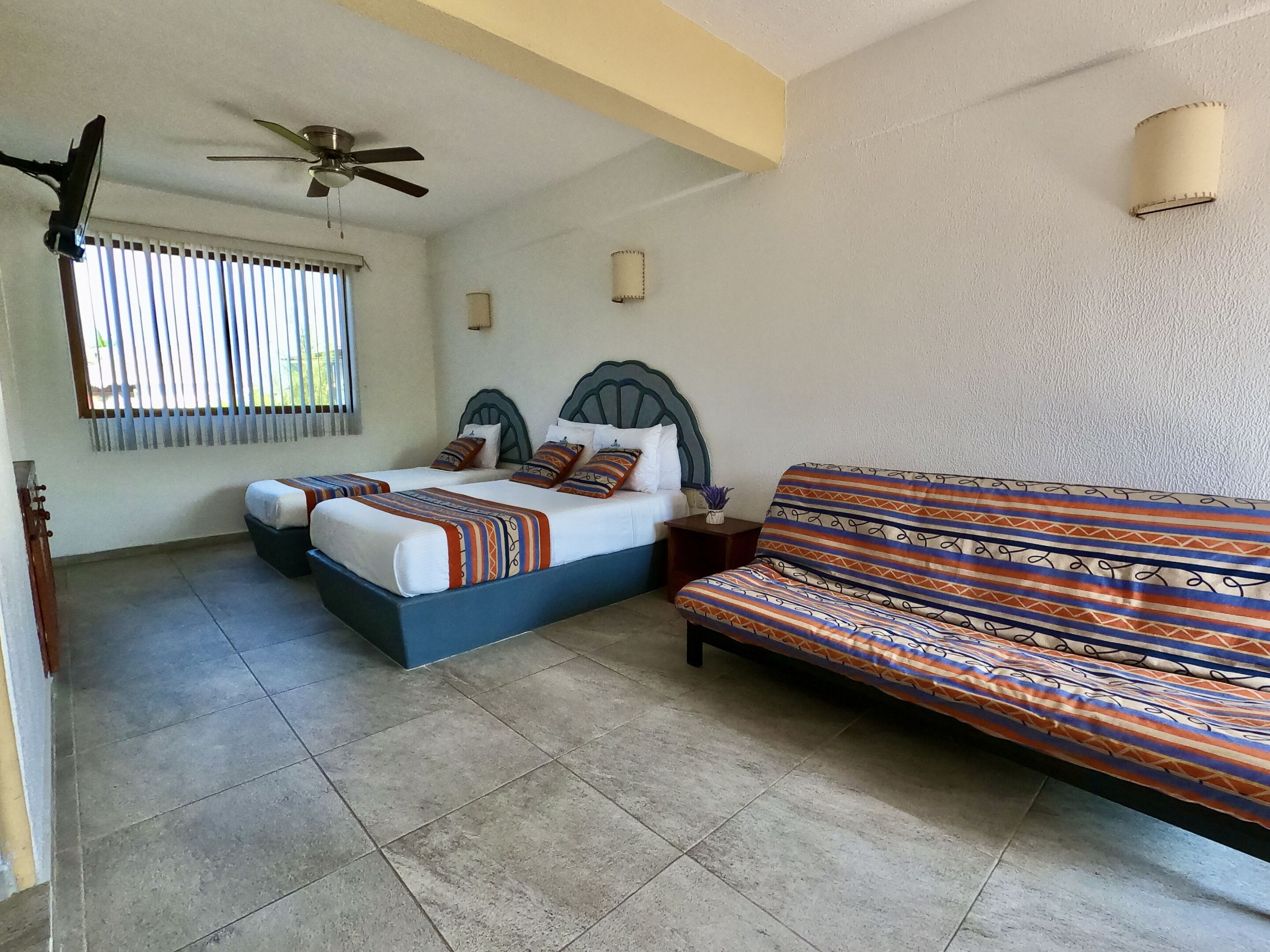 A bedroom with two beds, a sofa, a ceiling fan, and a window with blinds.