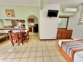 Una habitación con una cama, un escritorio, una silla, un televisor montado en la pared y una zona de cocina visible a través de un arco.