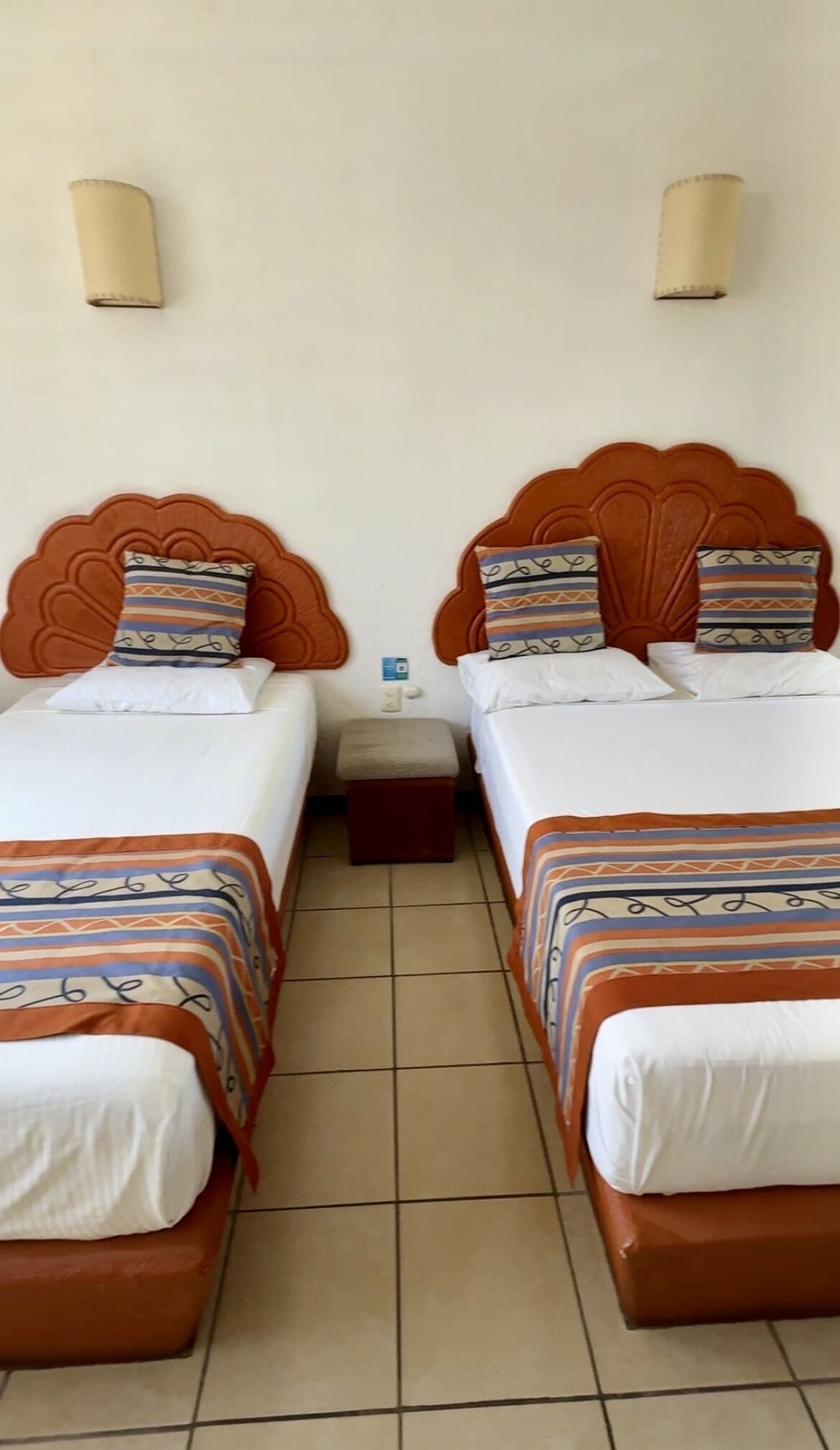 A hotel room with two beds, each with a patterned bedspread and matching pillows. There is a small bedside table with a lamp between the beds.