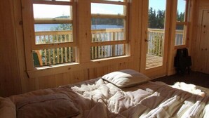 Een houten kamer met een bed, grote ramen en uitzicht op een houten terras en water.