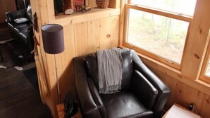 Een gezellig houten huisje van binnen, met een leren fauteuil, een klein tafeltje en een raam met uitzicht op de natuur.