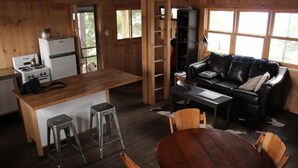 Een gezellig interieur van een chalet met een houten eettafel, een zwarte leren bank, een keuken met een fornuis en een koelkast, en een hond zichtbaar door een deur.