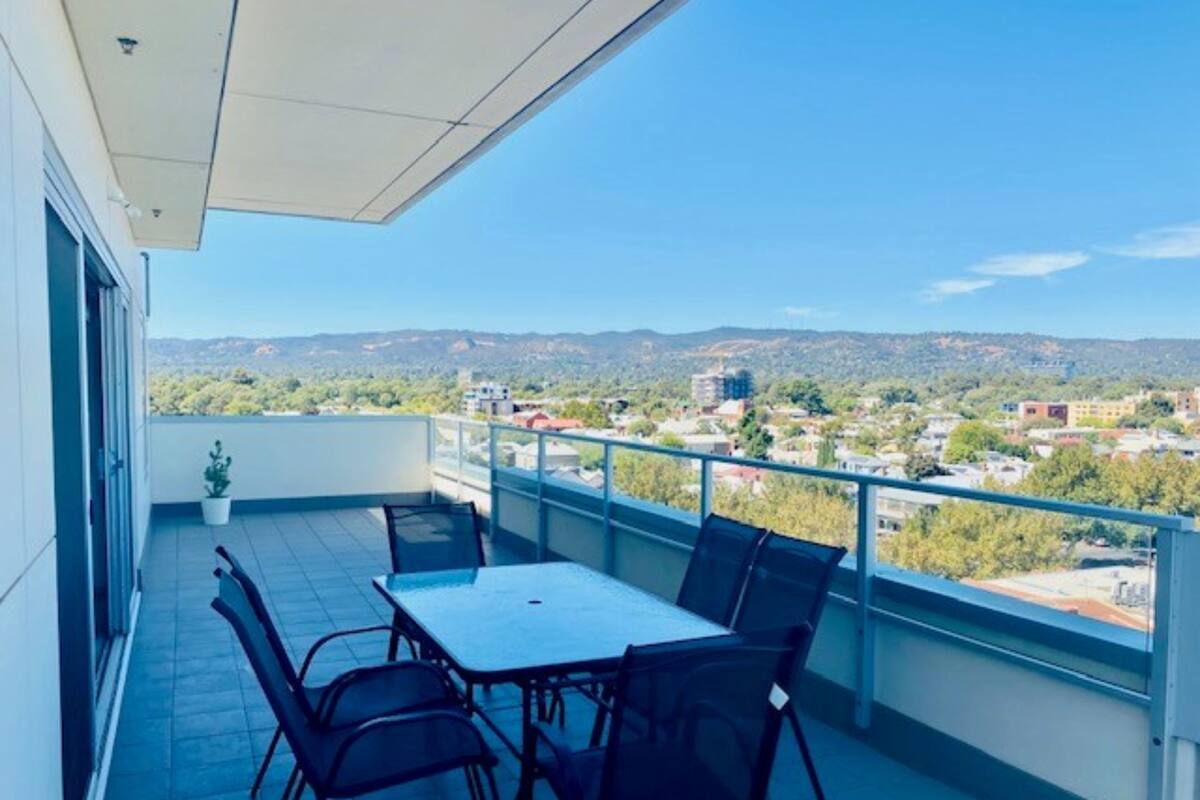 Un balcón con mesa y sillas, con vistas a un paisaje urbano y montañas.