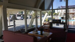 A hotel lobby with a red patterned sofa, a wooden coffee table, and a glass-top table with a vase. Large windows offer views of the street and a building with a pool. - Willow Tree Lodge (Fullerton)