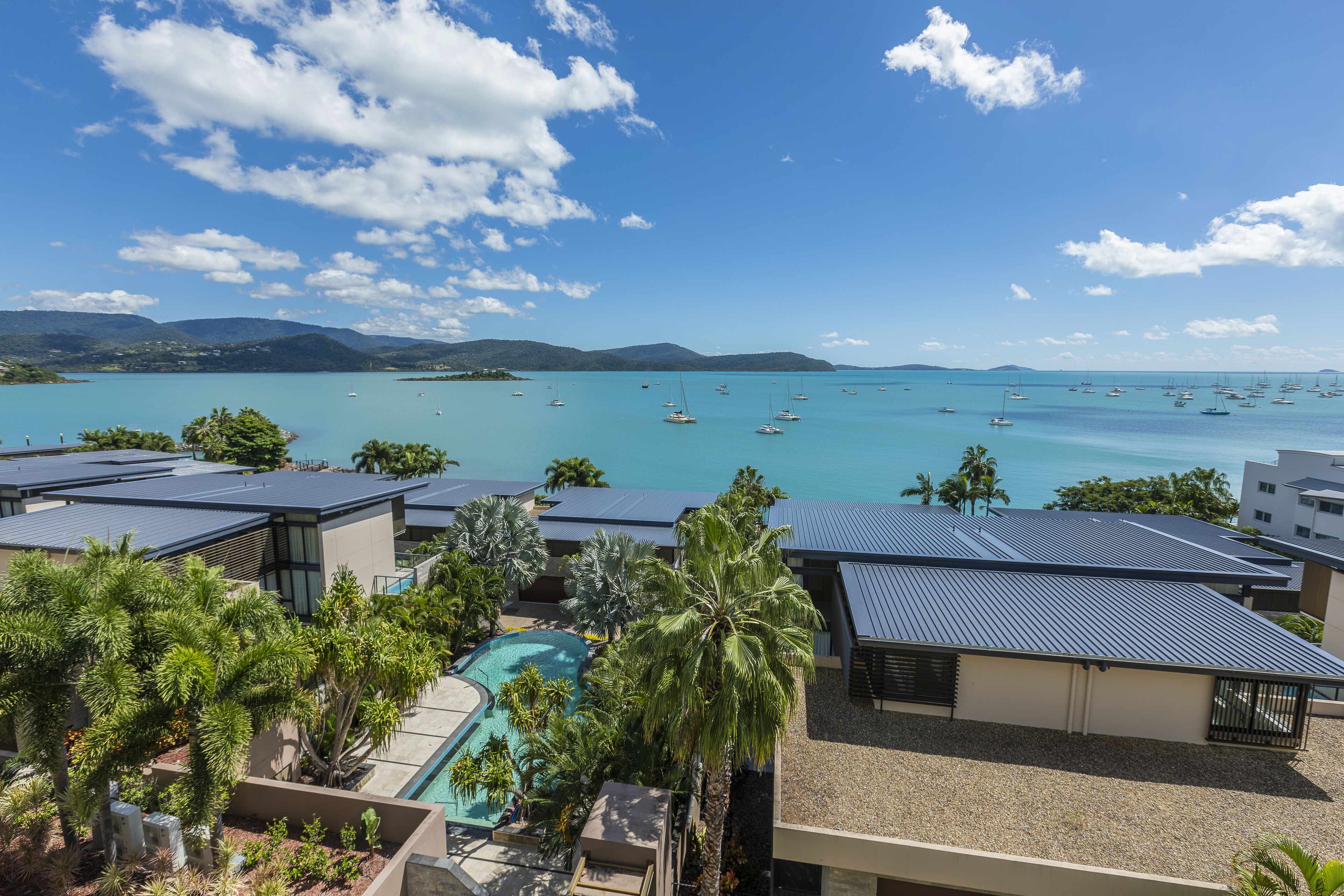 A resort with a pool, palm trees, and a view of the sea with boats.