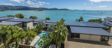 A resort with a pool, palm trees, and a view of the sea with boats.