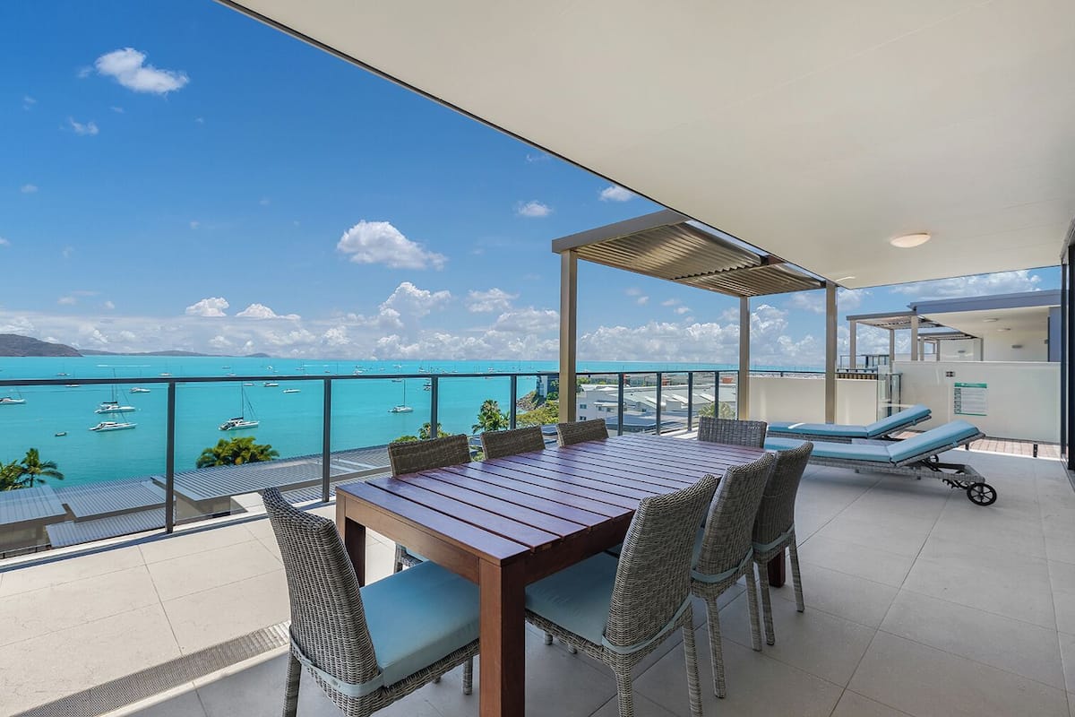 Un balcon avec une table en bois et des chaises en osier, donnant sur une mer turquoise où naviguent des bateaux.