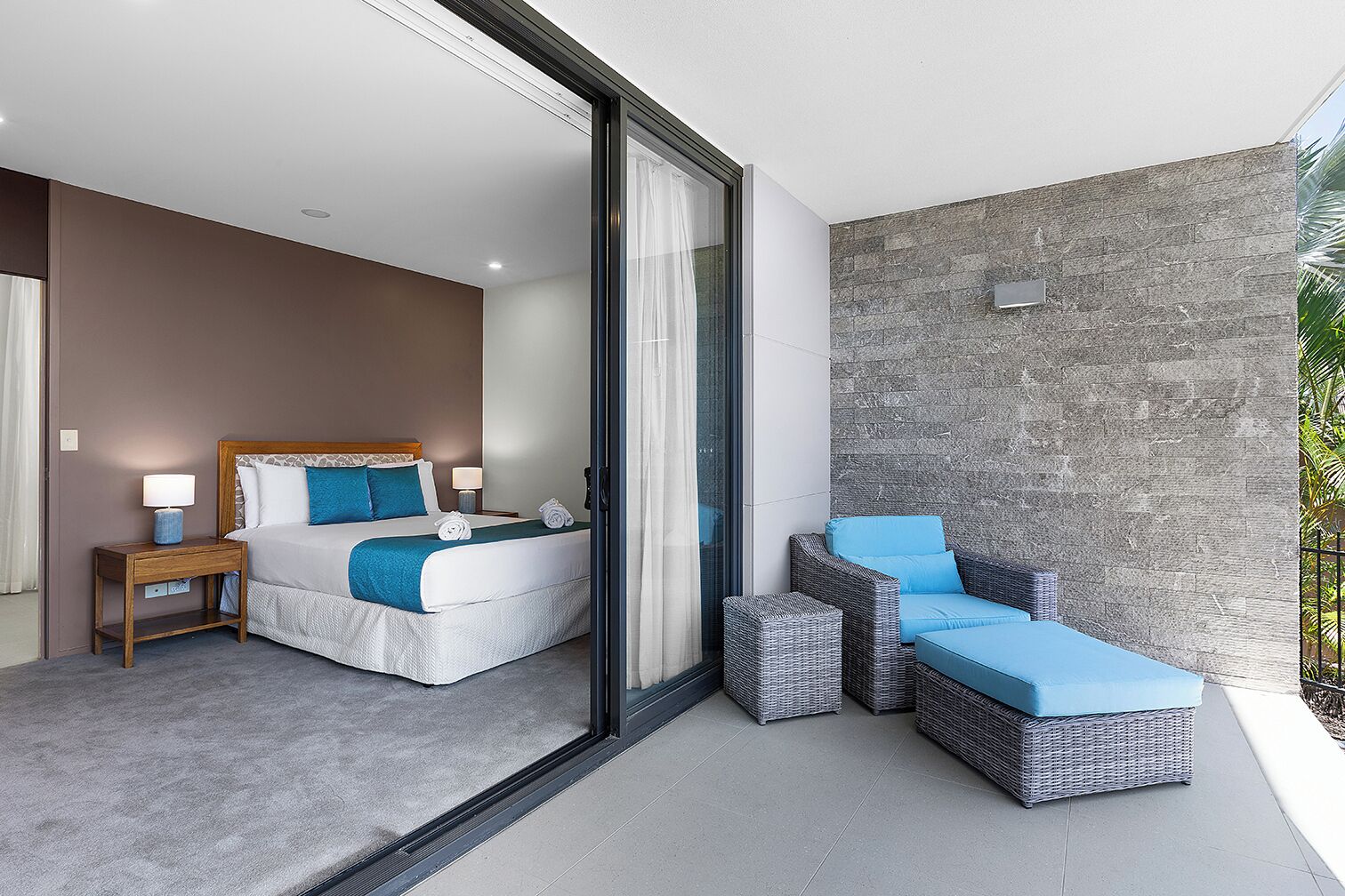 A modern hotel room with a large bed, a small bedside table, a nightstand, and a seating area with a wicker sofa and ottoman.