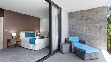 A modern hotel room with a large bed, a small bedside table, a nightstand, and a seating area with a wicker sofa and ottoman.
