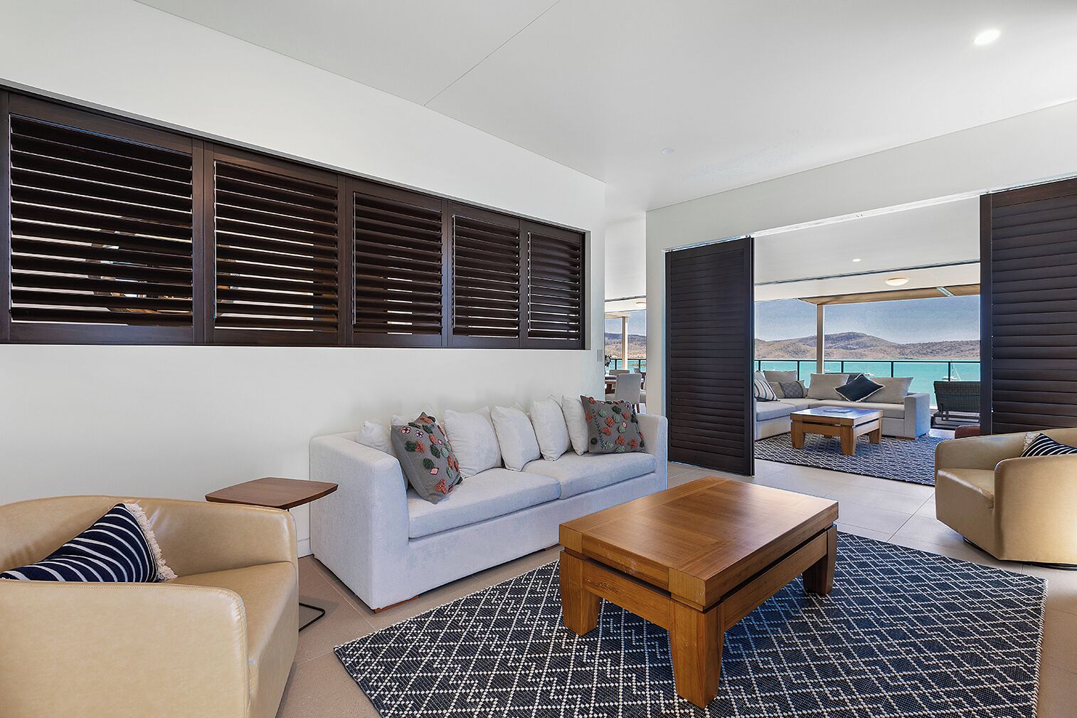 A modern living room with a sofa, coffee table, and two armchairs. A large window with blinds is visible, and a balcony with a view of the sea is in the background.
