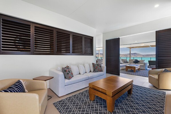 A modern living room with a sofa, coffee table, and two armchairs. A large window with blinds is visible, and a balcony with a view of the sea is in the background. - Mirage Whitsundays (Cannonvale)