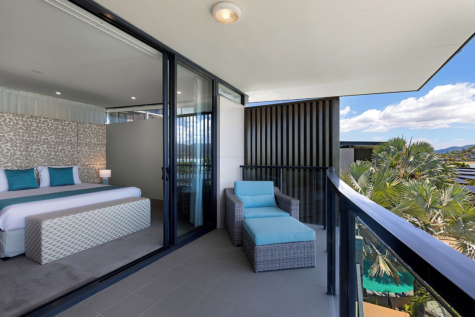 Une chambre moderne dotée d’un grand lit, d’un coin salon séparé avec une chaise longue, et d’un balcon offrant une vue sur des palmiers et un paysage lointain.