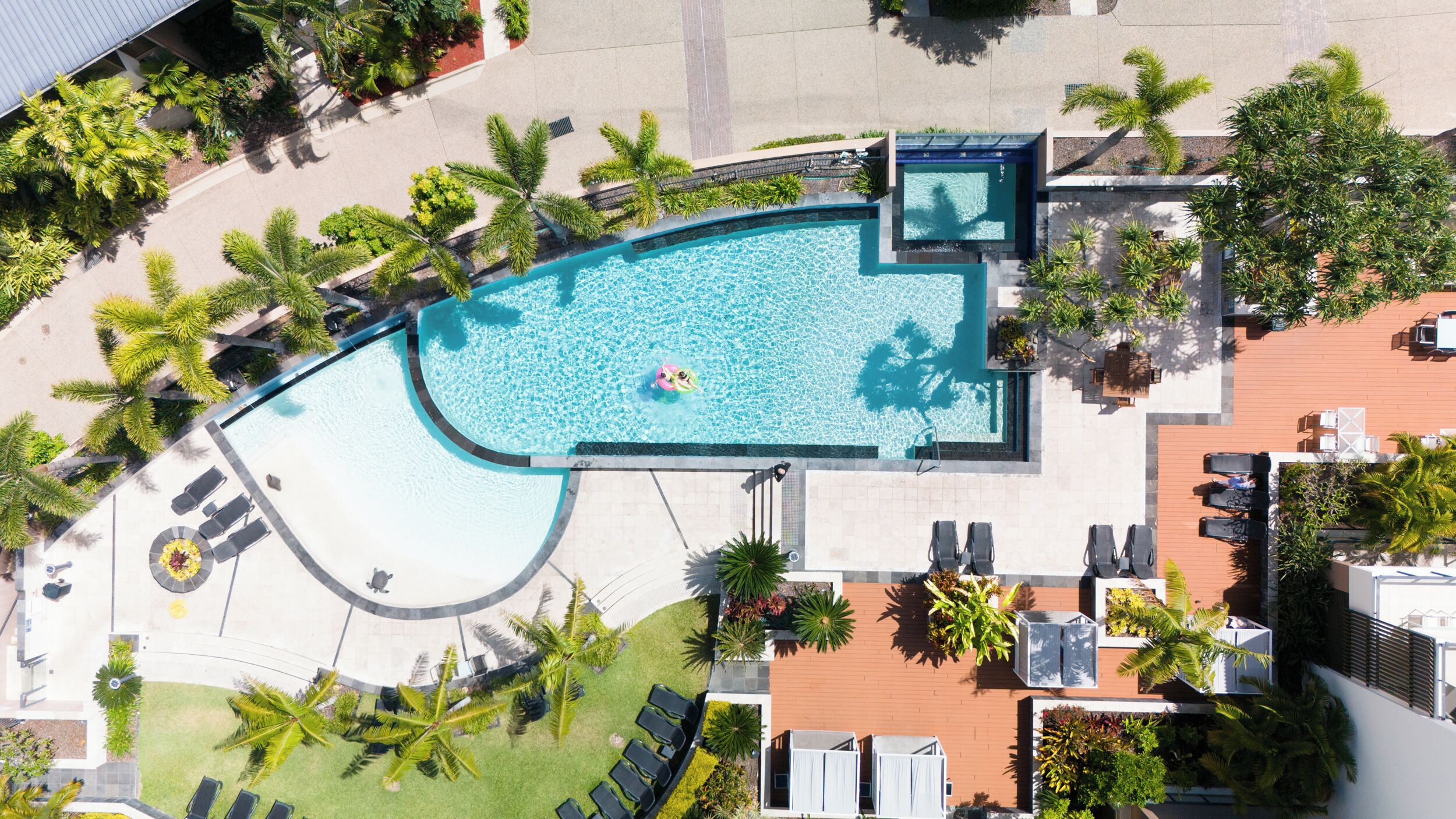 An aerial view of a resort with a large pool, sun loungers, and palm trees.