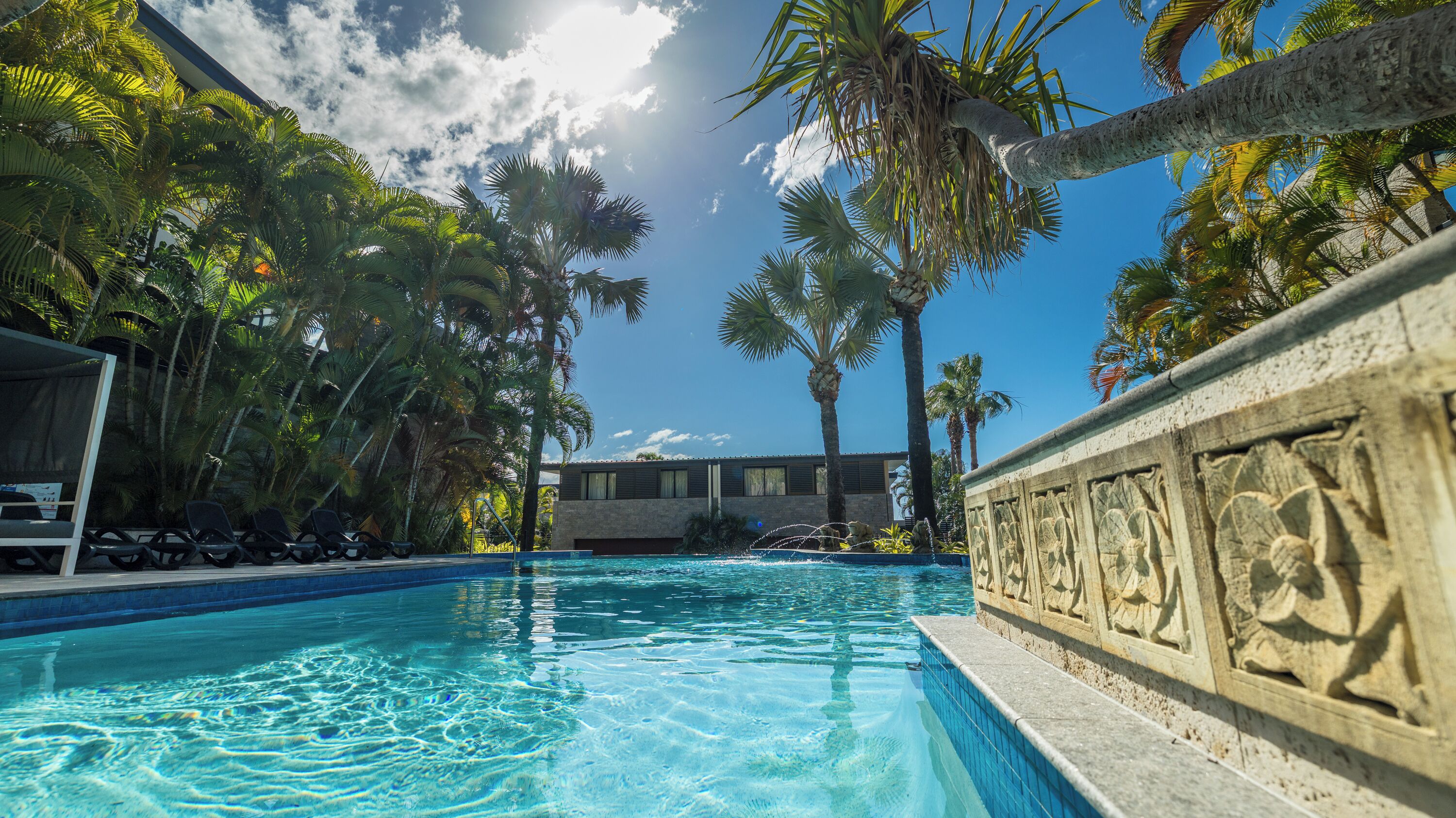 A swimming pool with clear blue water, surrounded by palm trees and a stone wall with carvings.
