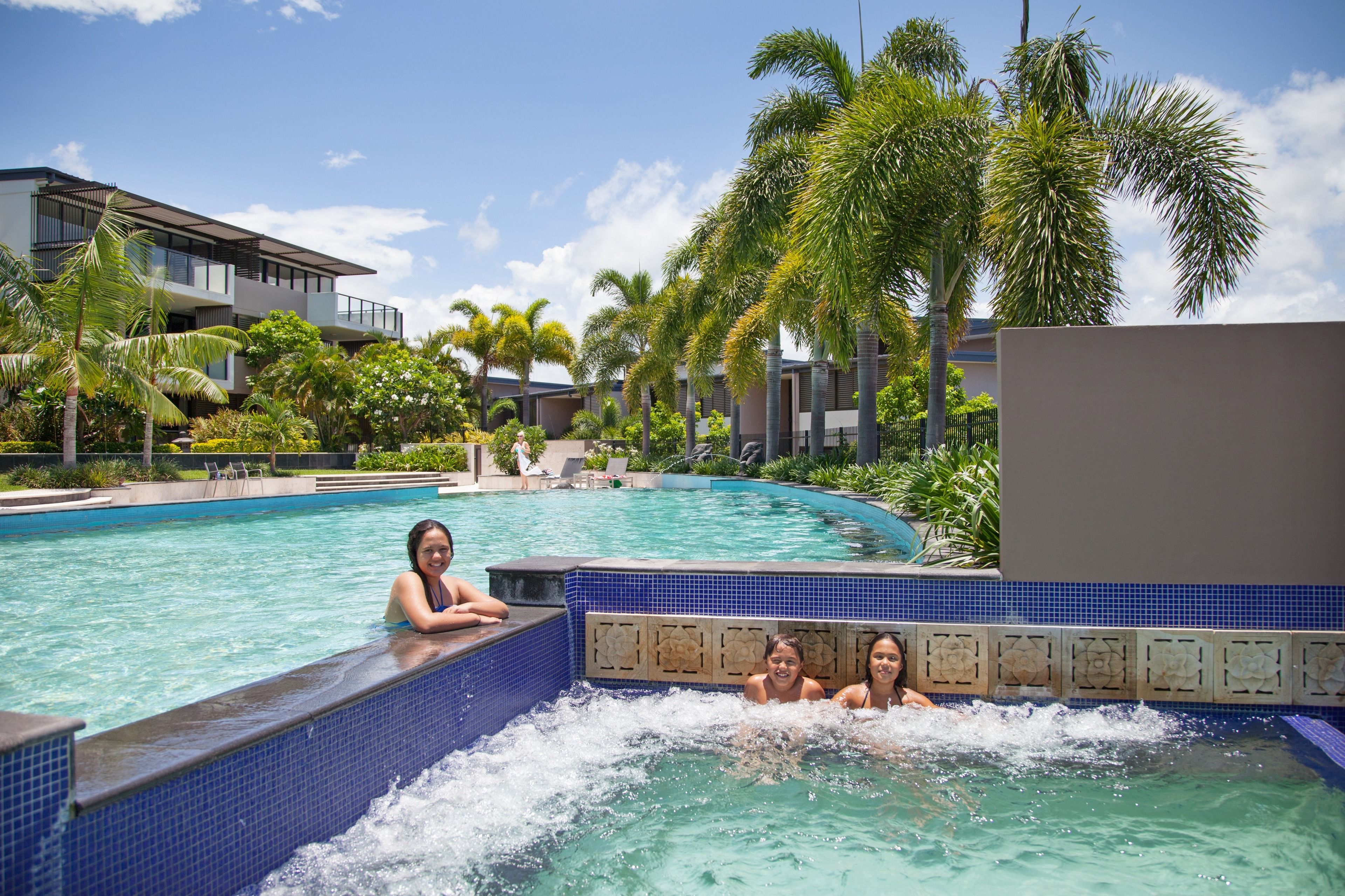 A swimming pool with a blue tiled edge, surrounded by palm trees and modern buildings.