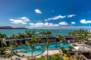 A resort with a large pool, palm trees, and a view of the ocean with boats.