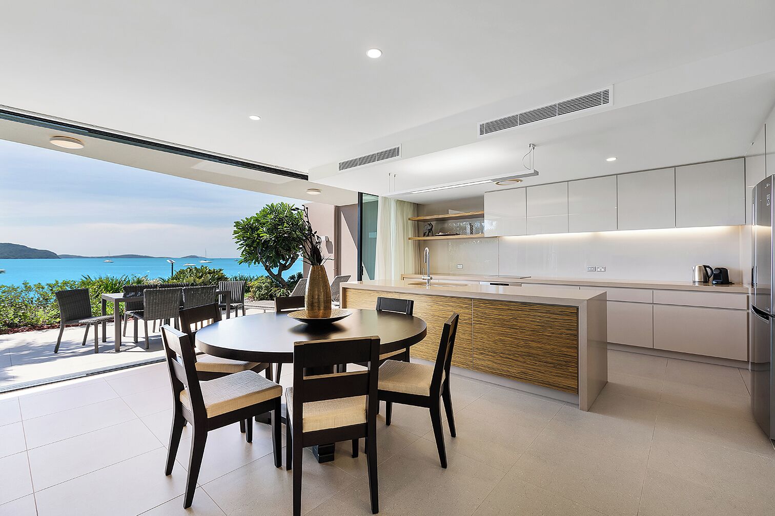 A modern kitchen with a view of the sea, featuring a dining area with chairs and a table, and a large island with a sink and cabinets.