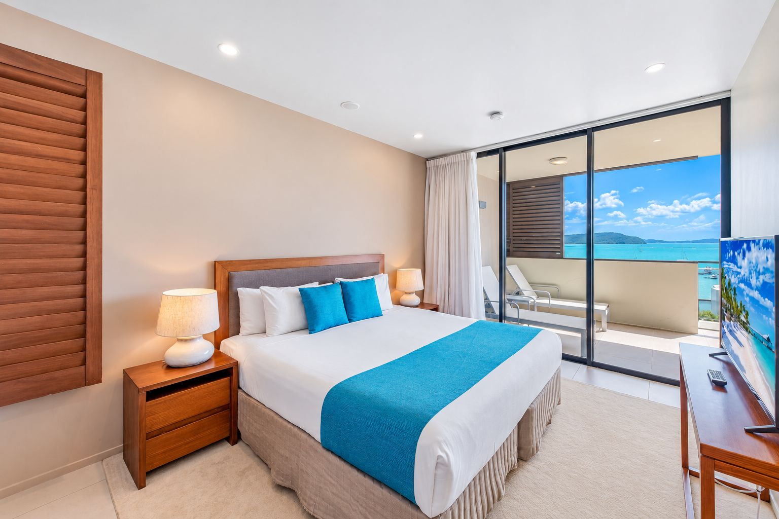 A hotel room with a bed, bedside lamps, a TV, and a balcony with a view of the sea.