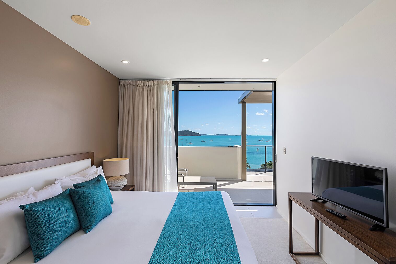 Quarto de hotel com uma cama, televisão, luminária e vista para o mar.