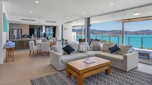 Un salon moderne avec un grand canapé, une table basse et un coin repas avec des chaises. De grandes fenêtres offrent une vue sur la mer et les montagnes.