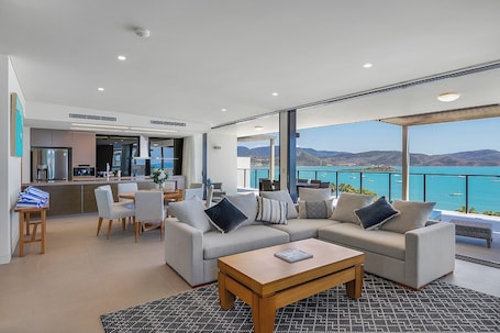 Una sala de estar moderna con un sofá grande, una mesa de centro y un comedor con sillas. Se ve el mar y las montañas a través de amplios ventanales.