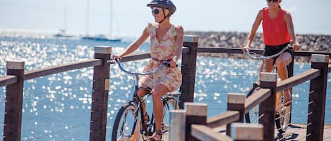 Deux personnes à vélo sur une jetée en bois, avec un plan d’eau et des bâtiments éloignés en arrière-plan.