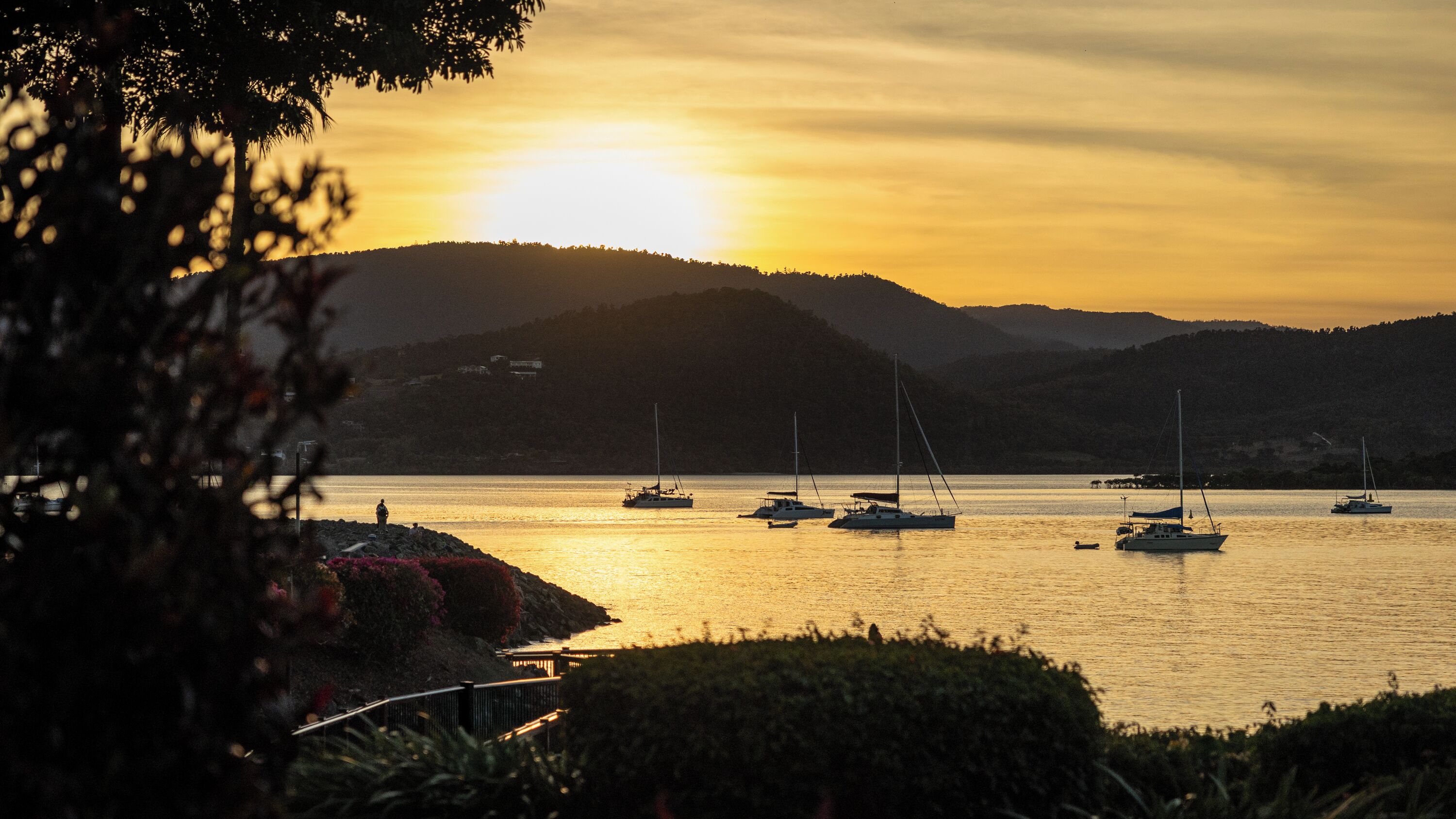A coastal scene with boats on calm water, a sunset, and a hilly landscape in the background.
