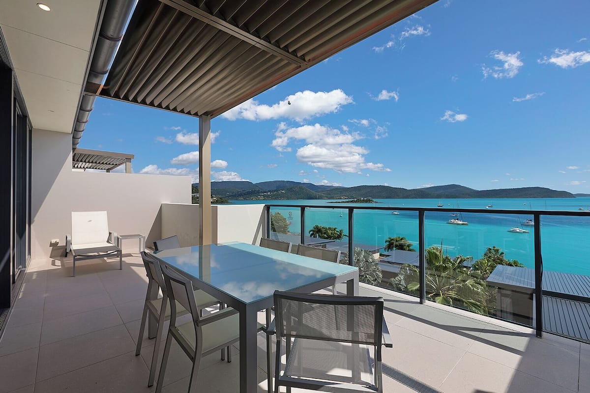 Un balcon avec une table et des chaises en verre, donnant sur une mer turquoise où l’on aperçoit des bateaux et un paysage montagneux.