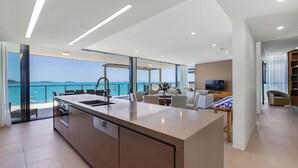 Une cuisine moderne dotée d’un grand îlot central, un coin repas offrant une vue sur la mer et un salon avec un téléviseur et des fauteuils.