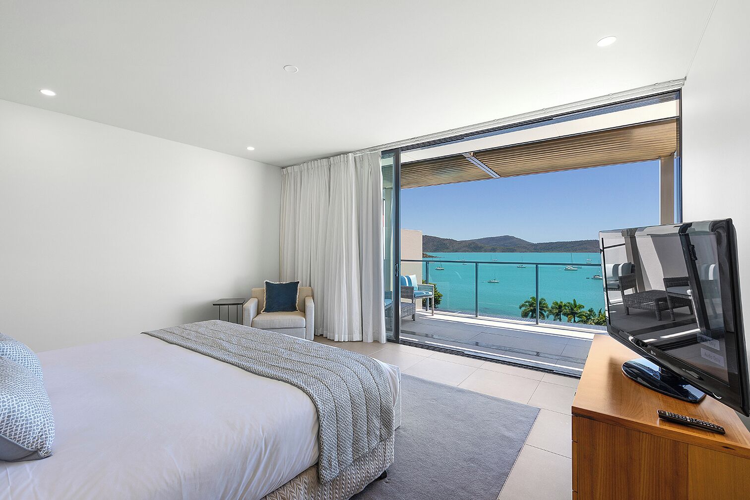 Una moderna camera d'albergo con un letto grande, un'area salotto con una poltrona e un tavolino, un balcone con vista sul mare e una TV a schermo piatto su un supporto in legno.