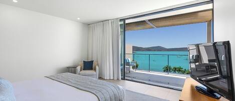 A modern hotel room with a large bed, a seating area with a chair and a small table, a balcony with a view of the sea, and a flat-screen TV on a wooden stand.
