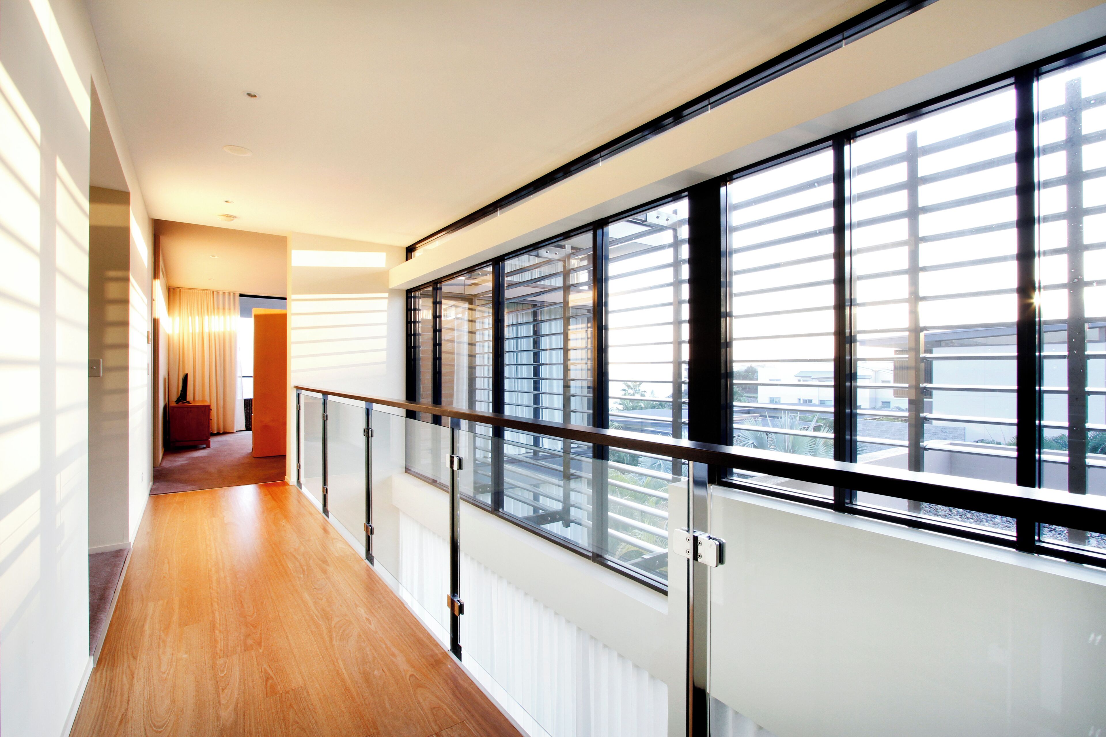 A modern hotel corridor with wooden flooring, a glass railing, and large windows offering a view of the cityscape.