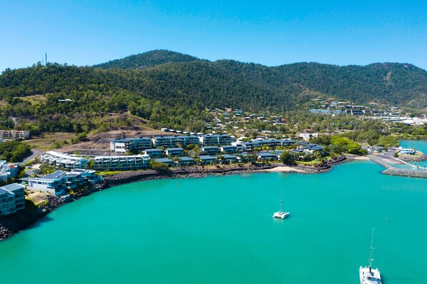An aerial view of a coastal resort with multiple buildings, a marina, and a backdrop of forested hills.