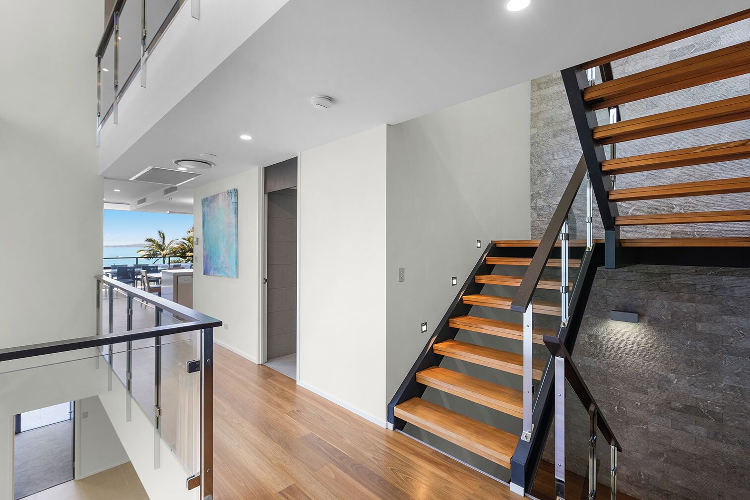 Un intérieur moderne avec un escalier en bois, des rampes en verre et une vue sur la mer.