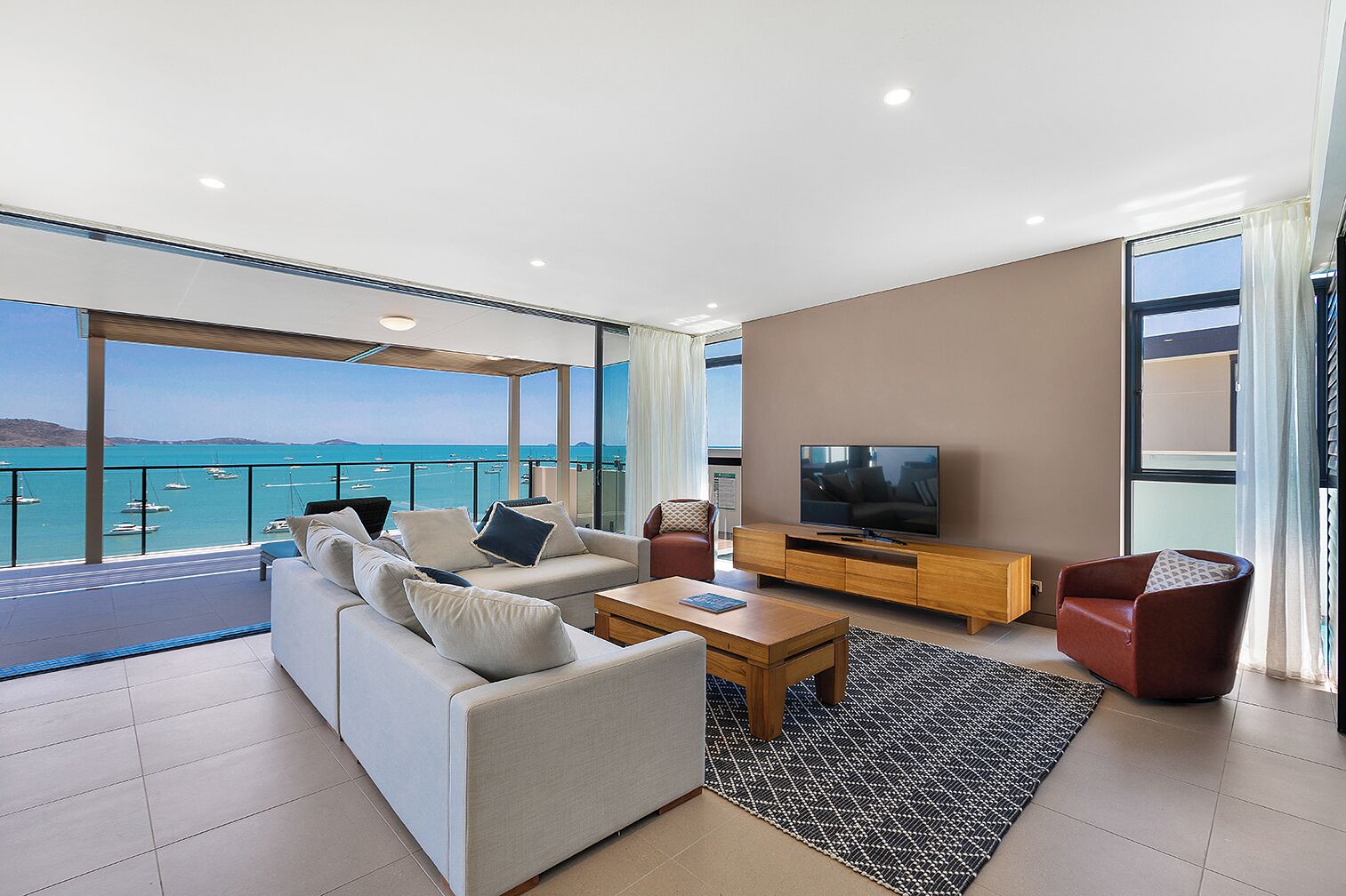 A modern living room with a large sofa, a coffee table, and a TV on a wooden stand. There is a balcony with a view of the sea and boats.