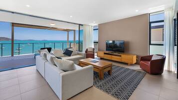 A modern living room with a large sofa, a coffee table, and a TV on a wooden stand. There is a balcony with a view of the sea and boats.